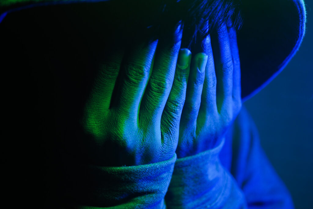Individual covers face with hands, surrounded by deep blue and green lighting.
