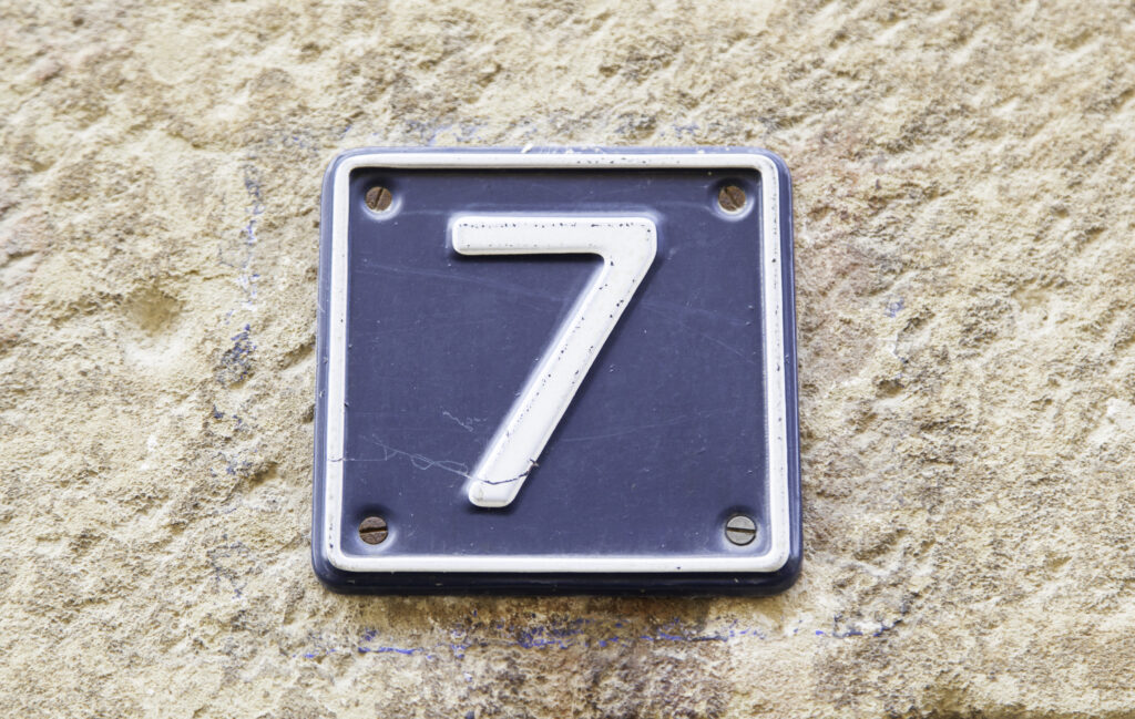 A close up shot of a number seven. The number seven is light gray on a blue square with gray trim. The square is mounted on a stone wall.