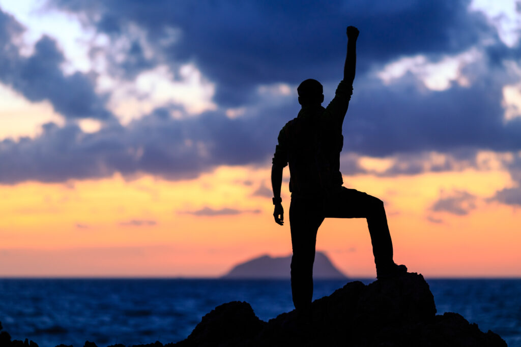 A silhouette shows an individual standing on a rock with one arm raised in a gesture of accomplishment, set against a beautiful sunset and ocean view. The sky is a blend of blues, oranges, and greys, indicating either sunrise or sunset. The water ripples in the foreground.